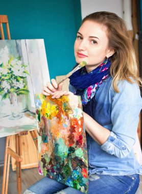 Elinde boya paleti olan bir kız sanatçı. Doğrudan kameraya bakıyor. Sanat stüdyosu