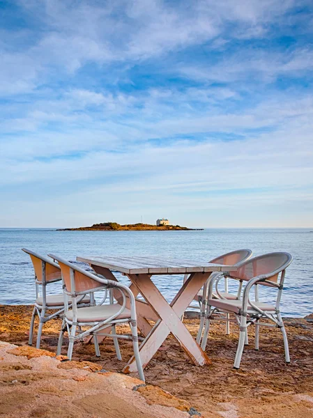 A romantic table with chairs on the island of Crete in Greece, Europe. Greek restaurant type of setup in front of the blue sea and an island with a greek chapel.