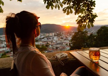 Genç kadın ile bardak bira batımını şehir Graz Avusturya üzerinden siting. Schlossberg Hill, saat kulesi ünlü turizm restorandan görüntülemek