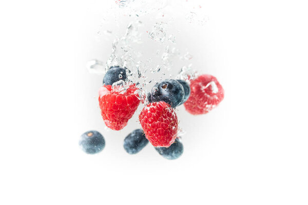 Blueberrys and raspberrys splashing into crystal clear water with air bubbles and sinking white background