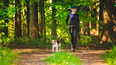 Sport girl is running with a dog (Beagle) at the spring sunny day on the forest path.