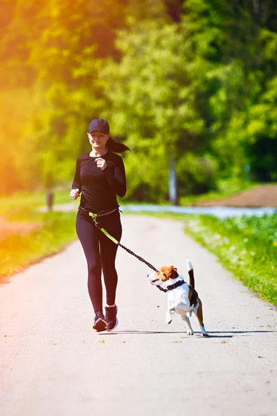 Sport girl is running with a dog (Beagle) on the rural road.