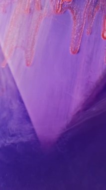 Dikey video. Boya damlası. Işıldayan damlalar. Mor üçgen üzerinde kayan turuncu simli damlalar renkli dumanlı su arkaplanı bulanıklaştırır.