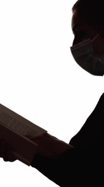 Dikey video. Karantina çalışması. Siluet okunuyor. Salgın hobisi. Yüz maskesi takmış, açık kitabı beyaz boş arkaplanda izole bir kadının koyu renk kontrast profili..