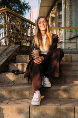 Kahve molası. Açık hava eğlencesi. Neşeli, rahat iş kadını merdivenlerde oturmuş kağıt bardakta sıcak içecekle eğleniyor..