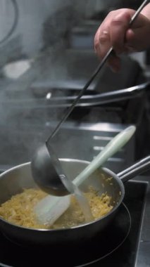 Dikey video. Pilav. Sıcak paella. Şef kızartma tavasına spatulaya kepçe suyu ekliyor. Kaynar su kaynayan yiyecekler.