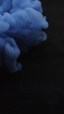 Dikey video. Duman boyası. Mistik duman. Mavi bulut spreyi siyaha döküldü. Suyun soyut arka planında renk üfleme bulanıklığı patlaması.