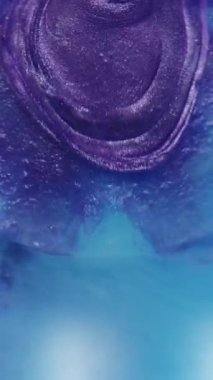 Dikey video. Işıldayan damlalar. Boya yayılmış. Mor parıltı damlaları bulanık elmas mavisi suyun soyut arka planına düşüyor.