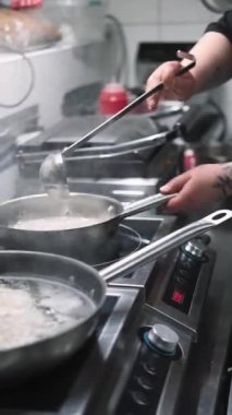 Dikey video. Güveç tabağı. Acı sos. Aşçı tavaya kepçe suyu ekliyor. Lokanta mutfağında kaynayan gurme yemekleri..