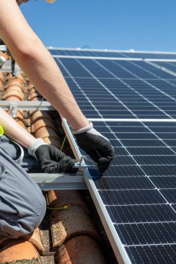 Güvenlik donanımlı bir işçi, parlak mavi gökyüzünün altındaki bir çatıya dikkatlice güneş panelleri yerleştiriyor. Görev, verimlilik ve istikrarı sağlamak için panelleri sıkıca güvence altına almayı içeriyor..