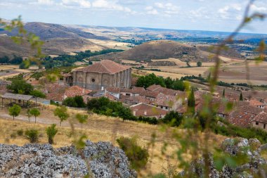 Guadalajara 'daki Atienza' ya bakan bu bakış açısı, bulutlu bir gökyüzünün altındaki yuvarlak tepeler ve yeşilliklerle çerçevelenmiş geniş bir kırsal alana kurulmuş tarihi binaları gözler önüne seriyor..