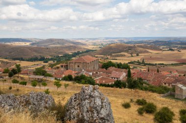 Guadalajara 'daki Atienza' ya bakan bu bakış açısı, bulutlu bir gökyüzünün altındaki yuvarlak tepeler ve yeşilliklerle çerçevelenmiş geniş bir kırsal alana kurulmuş tarihi binaları gözler önüne seriyor..