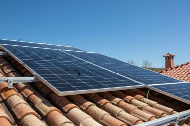 Güneş panelleri açık mavi bir gökyüzünün altında fayanslı bir çatıya monte edilir. Kurulumda konut kullanımı için çevre dostu enerji üretimi sergileniyor.