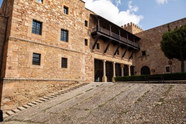Tarihi bir taş kale, verimli yeşillik ve parlak mavi gökyüzü ile çevrili karmaşık mimari ayrıntılar ve kuleler içerir ve gün boyunca ihtişamını sergiler..