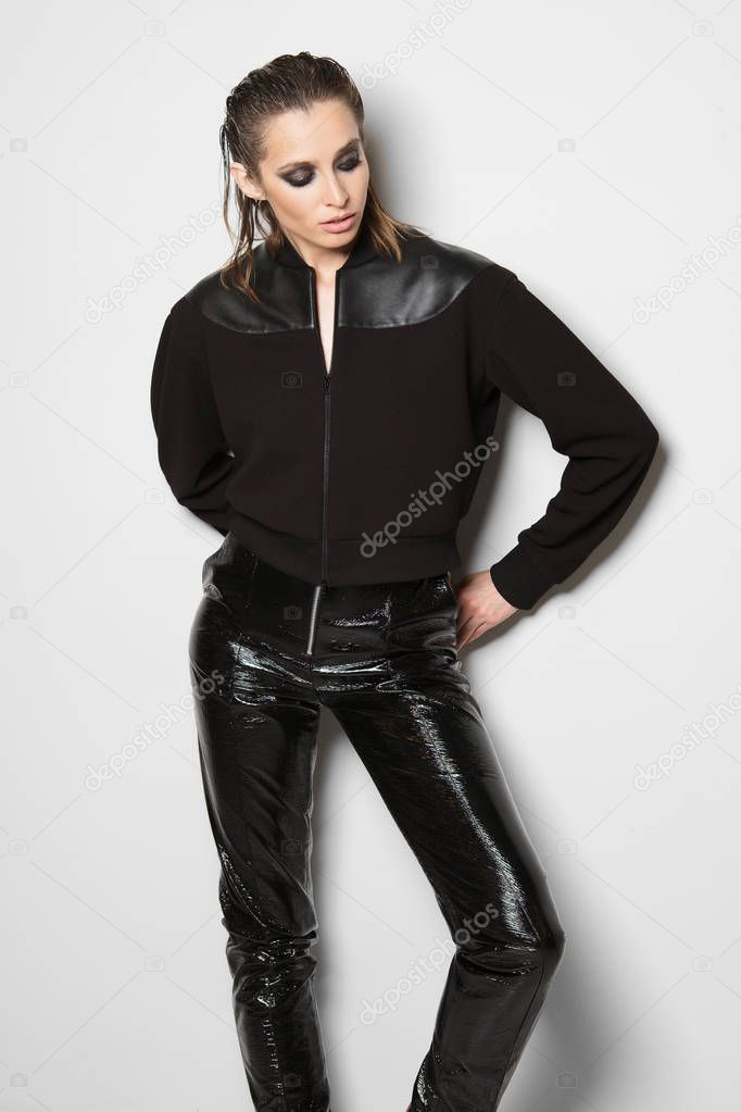 Portrait of caucasian woman in zipper sweater with leather inserts and faux leather pants posing against white wall.