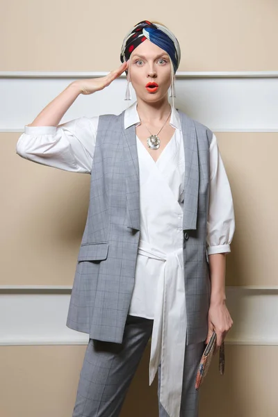 Surprised lady in sleeveless jacket, long asymmetric  shirt,  pants and turban posing with arm near head