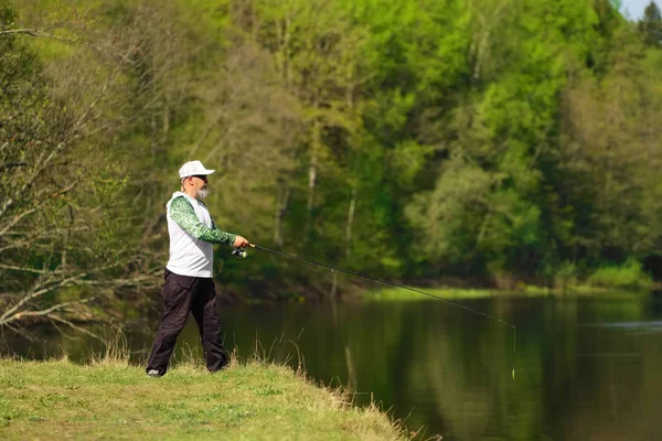 Pescatore con una canna da spinning cattura pesce su un fiume in soleggiata giornata estiva con alberi verdi sullo sfondo. Attività all'aperto nel fine settimana. Foto con bassa profondità di campo scattata a grande apertura . — Foto Stock