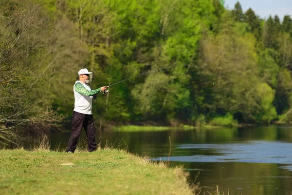 Fischer mit einer Spinnrute, die an einem sonnigen Sommertag auf einem Fluss Fische fängt, mit grünen Bäumen im Hintergrund. Aktivitäten im Freien am Wochenende. Foto mit geringer Schärfentiefe bei weit geöffneter Blende. — Stockfoto