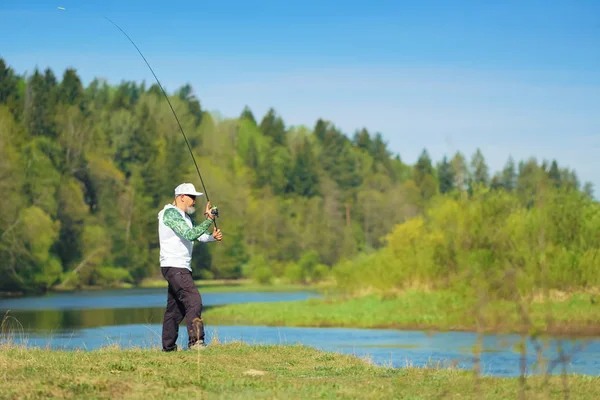 Рибалка з обертовим стрижнем ловить рибу на річці в сонячний літній день з зеленими деревами на фоні. Активність на відкритих вихідних. Фотографія з неглибокою глибиною різкості, зроблена на широкому відкритому отворі . — стокове фото