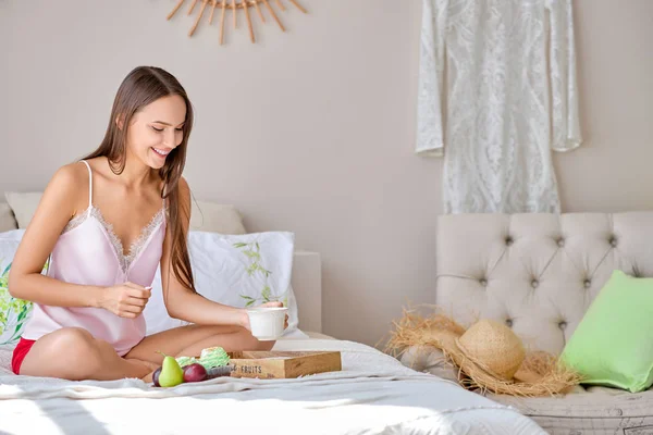 Boldog fiatal hölgy vesz csésze teát evés közben az ágyban — Stock Fotó