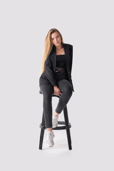 Retrato de comprimento total de bela jovem sentada em cadeira alta em jeans pretos e jaqueta sobre fundo cinza — Fotografia de Stock