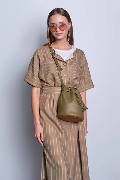 Trendige Dame mit großer Sonnenbrille trägt gestreiftes Olivkleid mit Lederhandtasche vor grauem Hintergrund — Stockfoto