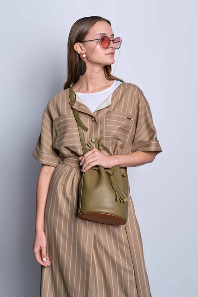 Dame à la mode dans de grandes lunettes de soleil portant une robe d'olive rayée avec un sac à main en cuir posant sur fond gris — Photo