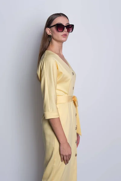 Модель в больших солнцезащитных очках в жёлтом платье с пуговицами на сером фоне — стоковое фото