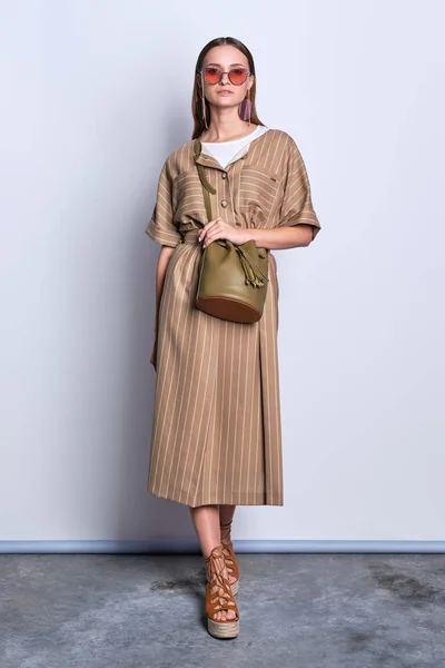 Dame à la mode dans de grandes lunettes de soleil portant une robe d'olive rayée avec un sac à main en cuir posant sur fond gris — Photo