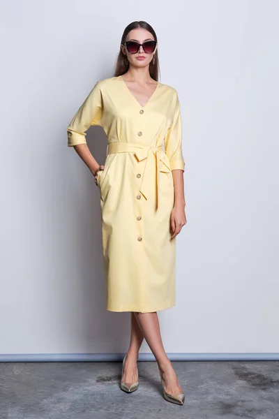 Fashion model in big sunglasses wearing yellow dress with buttons posing over gray background