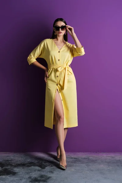 Fashion model in big sunglasses wearing yellow dress with unfastened buttons near purple wall