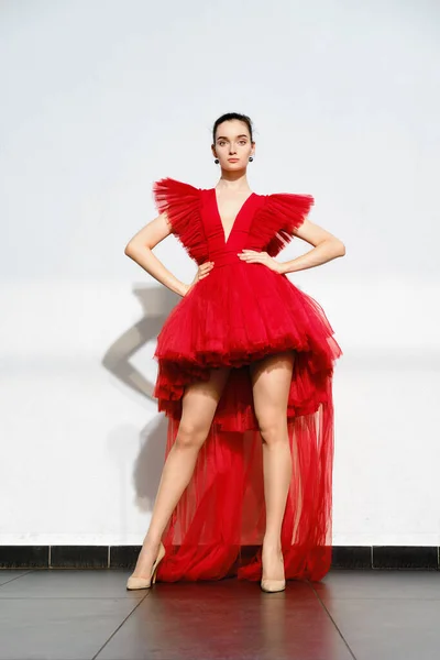 Beautiful girl in short red tulle dress with long tail posing under hard light