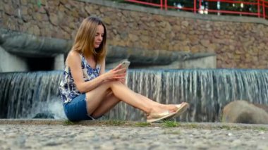 Şehir parkında, şelalenin yanındaki kaldırım taşlı rıhtımda oturan ve cep telefonundan haber okuyan bir kızın görüntüsü düşük.