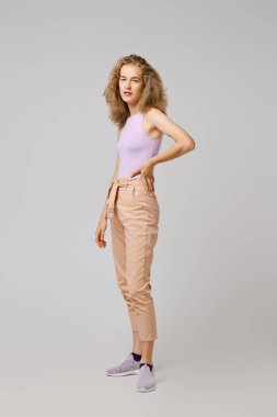 Sarışın, dağınık saçlı, kolsuz tişörtlü, pantolonlu, gri arka planda poz veren güzel kız.