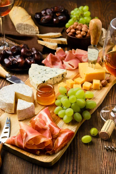Peynir tabağında prosciutto, üzüm, bal, hurma, kraker, fındık ve şarap var.