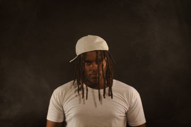 Rastalı ve beyaz şapkalı yakışıklı bir genç stüdyoda fotoğraf çekimi sırasında kameraya poz veriyor. Dumanlı bir arkaplanda izole edilmiş.