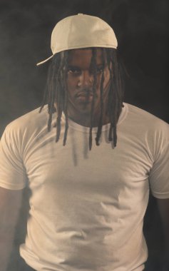 Rastalı ve beyaz şapkalı yakışıklı bir genç stüdyoda fotoğraf çekimi sırasında kameraya poz veriyor. Dumanlı bir arkaplanda izole edilmiş.