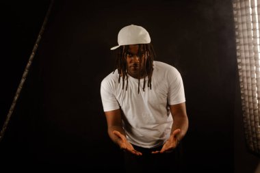 Rastalı ve beyaz şapkalı yakışıklı bir genç stüdyoda fotoğraf çekimi sırasında kameraya poz veriyor. Dumanlı bir arkaplanda izole edilmiş.