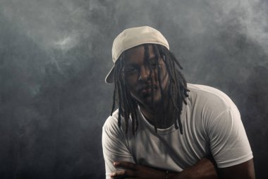 Rastalı, şapkalı ve beyaz gömlekli bir adam stüdyoda dumanlı bir arka planda kameraya poz veriyor. Kendinden emin ve ciddi bir genç adam..