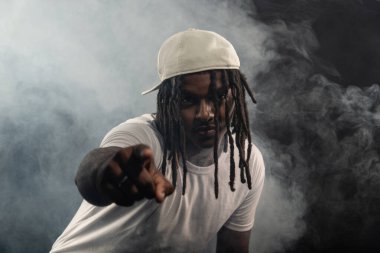 Rastalı, şapkalı ve beyaz gömlekli bir adam stüdyoda dumanlı bir arka planda kameraya poz veriyor. Kendinden emin ve ciddi bir genç adam..