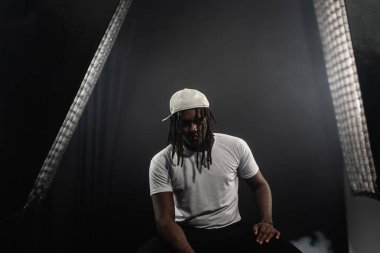 Rastalı, şapkalı ve beyaz gömlekli bir adam stüdyoda dumanlı bir arka planda kameraya poz veriyor. Kendinden emin ve ciddi bir genç adam..