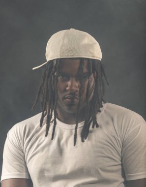 Rastalı, şapkalı ve beyaz gömlekli bir adam stüdyoda dumanlı bir arka planda kameraya poz veriyor. Kendinden emin ve ciddi bir genç adam..