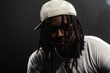 Rastalı, şapkalı ve beyaz gömlekli bir adam stüdyoda dumanlı bir arka planda kameraya poz veriyor. Kendinden emin ve ciddi bir genç adam..