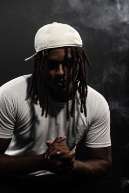 Rastalı, şapkalı ve beyaz gömlekli bir adam stüdyoda dumanlı bir arka planda kameraya poz veriyor. Kendinden emin ve ciddi bir genç adam..