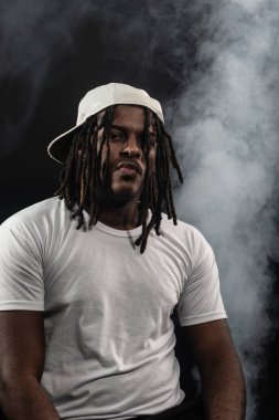 Rastalı, şapkalı ve beyaz gömlekli bir adam stüdyoda dumanlı bir arka planda kameraya poz veriyor. Kendinden emin ve ciddi bir genç adam..