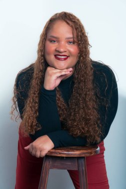 Kıvırcık saçlı, kırmızı pantolonlu, çenesi üstünde siyah gömlekli, beyaz arka planda kameraya bakan güzel bir kadın portresi..