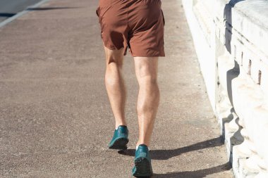 Bir insanın alt kısmı ve gölgesi, güçlü güneş ışığı altında sahil boyunca koşuyor. Sağlıklı spor pratiği. Brezilya.