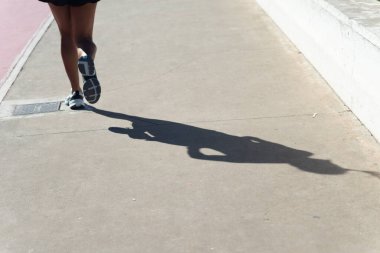 Bir insanın alt kısmı ve gölgesi, güçlü güneş ışığı altında sahil boyunca koşuyor. Sağlıklı spor pratiği. Brezilya.