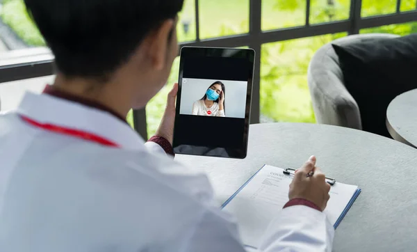 Doktorun bakış açısı hastayla iletişim kuruyor. Tabletlerle maske takıyor. Doktorlar, sosyal mesafeyi korumak için hastalarla video konferansı yoluyla iletişim kurmak için teletıp teknolojisini kullanıyorlar.