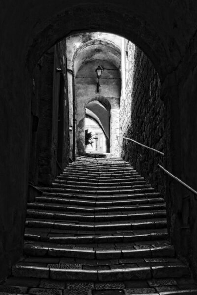 Suggestive covered staircase that climbs towards arches that cross where it ends. A ray of light penetrates between two arches and is reflected on the dark stone steps.